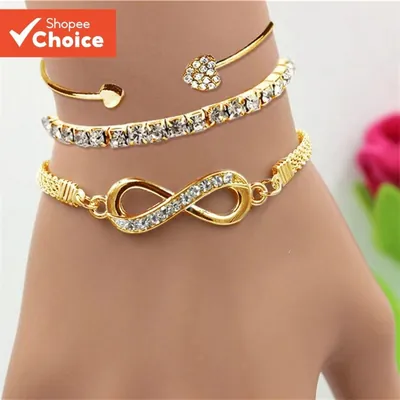 Pulso com três pulseiras douradas: bracelete com pontas de coração, riviera de pedras claras e corrente com infinito