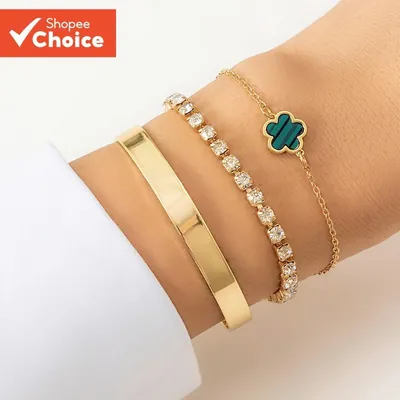 Três pulseiras douradas no pulso: bracelete liso, pulseira de strass e corrente com flor verde