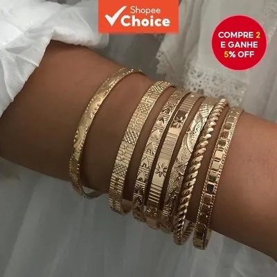 Braço com sete braceletes dourados finos de texturas diferentes, sobre blusa branca de renda