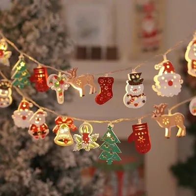 Cordão de luzes com pingentes acesos de Papai Noel, rena, boneco de neve, meia, sino e pinheiro diante de árvore nevada