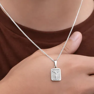 Homem de camiseta marrom usando corrente prateada fina com pingente retangular de Cristo em relevo apoiado no polegar