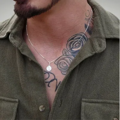 Homem de barba e camisa verde-oliva aberta usando corrente fina prateada com medalha redonda no peito tatuado