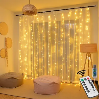 Cortina de fios de LED amarelo atrás de voal branco numa sala com pufes, luminária de tripé e controle remoto