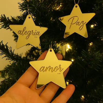 Três estrelas de acrílico dourado espelhado com as palavras alegria, paz e amor, penduradas numa árvore com luzes