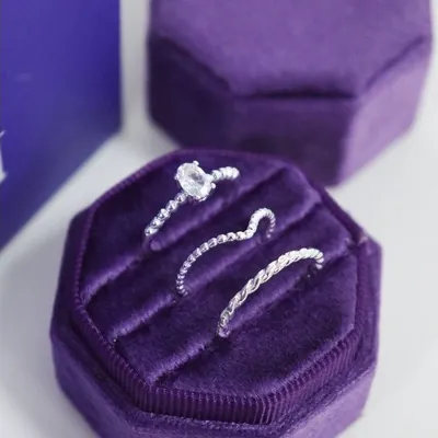 Três anéis finos prateados, um com pedra oval clara, encaixados em caixa octogonal de veludo roxo