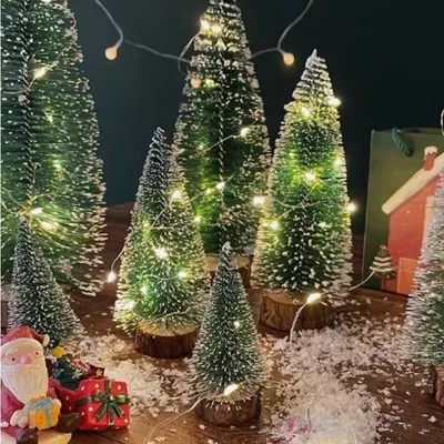Mini pinheiros nevados de alturas variadas sobre bases de tronco, com neve artificial e fio de luzinhas sobre mesa