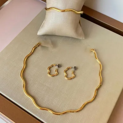 Choker rígida, pulseira e par de brincos de argola dourados em formato ondulado, expostos em caixa bege