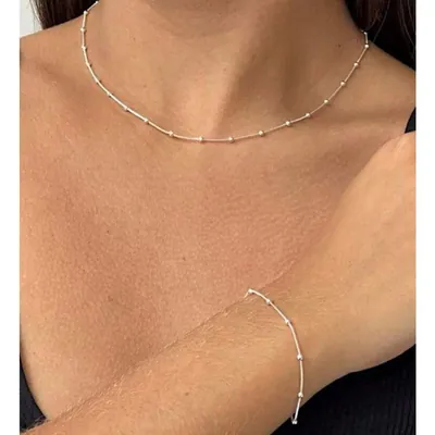 Colar curto e pulseira de fio fino prateado com bolinhas espaçadas, usados no pescoço e no pulso de uma mulher