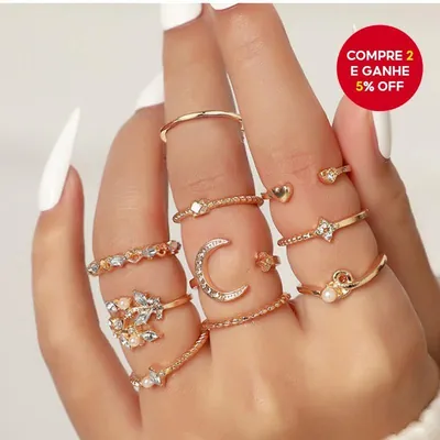 Mão feminina de unhas brancas usando vários anéis dourados finos com lua, estrela, coração, folhas e contas peroladas