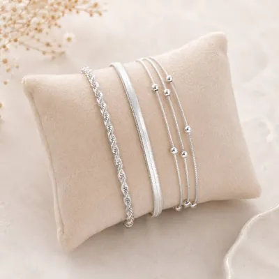 Pulseiras prateadas de cordão baiano, fio liso e correntes finas com bolinhas apoiadas numa almofada bege