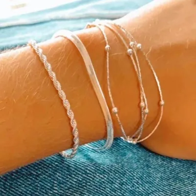 Pulso bronzeado sobre jeans com três pulseiras prateadas: cordão torcido, fita lisa achatada e fios finos com bolinhas