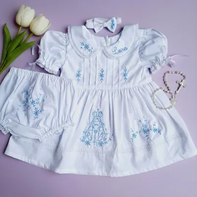 Vestido branco de bebê com bordado azul de Nossa Senhora, gola com renda, calcinha e faixa de laço sobre fundo lilás