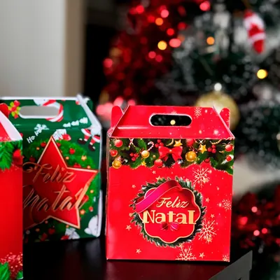Caixinhas de papel em forma de maleta, vermelha com selo Feliz Natal e verde com estrela, diante de árvore com luzes