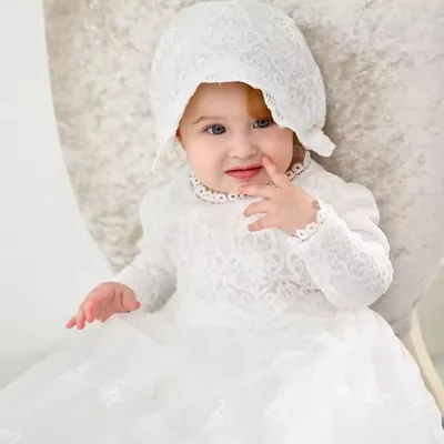Bebê sentada em poltrona clara usando mandrião branco de renda de manga longa e touca de renda amarrada