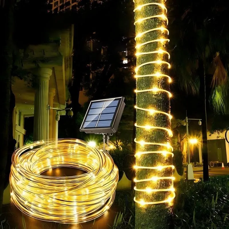 Mangueira de LED amarelada enrolada em espiral num tronco de árvore e em rolo no chão, com painel solar em estaca