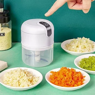 Mini processador branco com copo transparente e botão no topo, cercado de pratos com alho, cenoura e verdura picados
