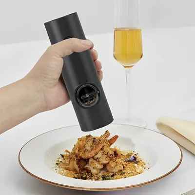 Mão segurando moedor elétrico preto cilíndrico sobre prato de camarões, com taça de vinho branco ao fundo