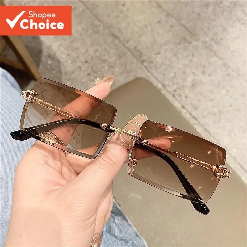 Mão segurando óculos de sol retangulares sem aro com lentes marrons em degradê e hastes douradas