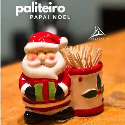 Paliteiro de Papai Noel com presente verde ao lado de copinho branco com azevinho, cheio de palitos, sobre balcão de madeira