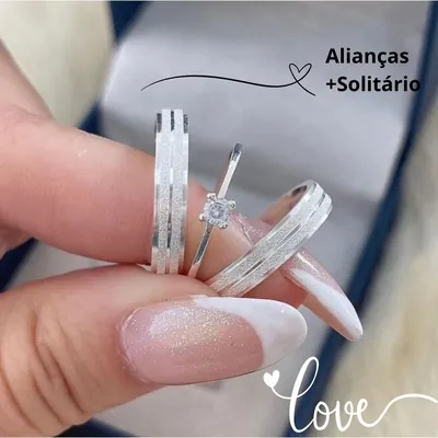 Duas alianças prateadas com faixas foscas diamantadas e um anel solitário de pedra clara presos entre dedos de mão feminina