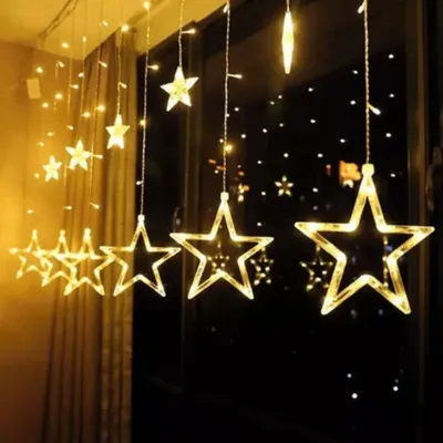 Cortina de luzes amareladas com fios pendentes e estrelas de LED de tamanhos variados diante de uma janela à noite