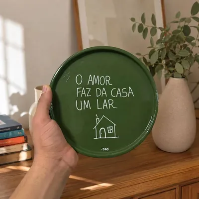 Mão segurando prato de argila verde esmaltado com a frase O amor faz da casa um lar e desenho de casinha em branco
