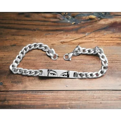 Pulseira prateada de elos grossos com placa central gravada com olhos e sobrancelhas em preto, sobre madeira rústica