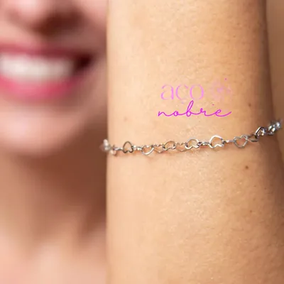 Pulso com pulseira prateada de elos em formato de coração vazado; ao fundo, rosto sorridente desfocado