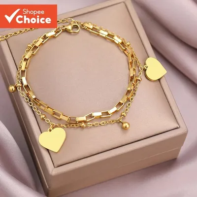 Pulseira dupla dourada com elos retangulares, corrente de bolinhas e dois corações, sobre caixa bege e tecido rosa