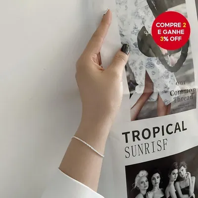 Pulso fino com pulseira prateada delicada e cintilante, mão de unhas escuras apoiada em pôster de revista na parede