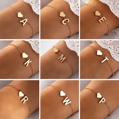 Nove pulsos com pulseira de corrente fina dourada, cada uma com um coração liso e uma letra maiúscula diferente