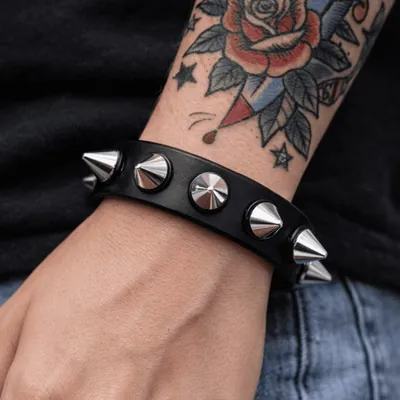 Pulseira preta com uma fileira de spikes cônicos prateados no pulso de pessoa com tatuagem de rosa no antebraço