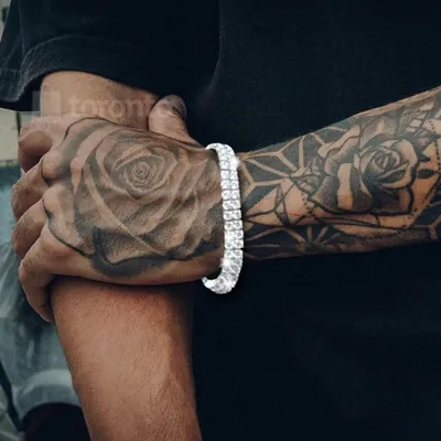 Pulso masculino tatuado com rosas usando pulseira prateada com duas fileiras de pedras brancas, sobre camiseta preta
