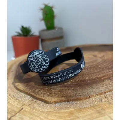 Pulseira de silicone preta com medalha de São Bento e oração em letras brancas, sobre tora de madeira com cactos ao fundo