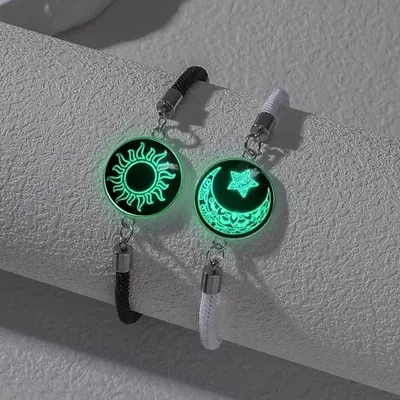 Duas pulseiras de cordão, uma preta com medalha de sol e uma branca com lua e estrela, brilhando em verde