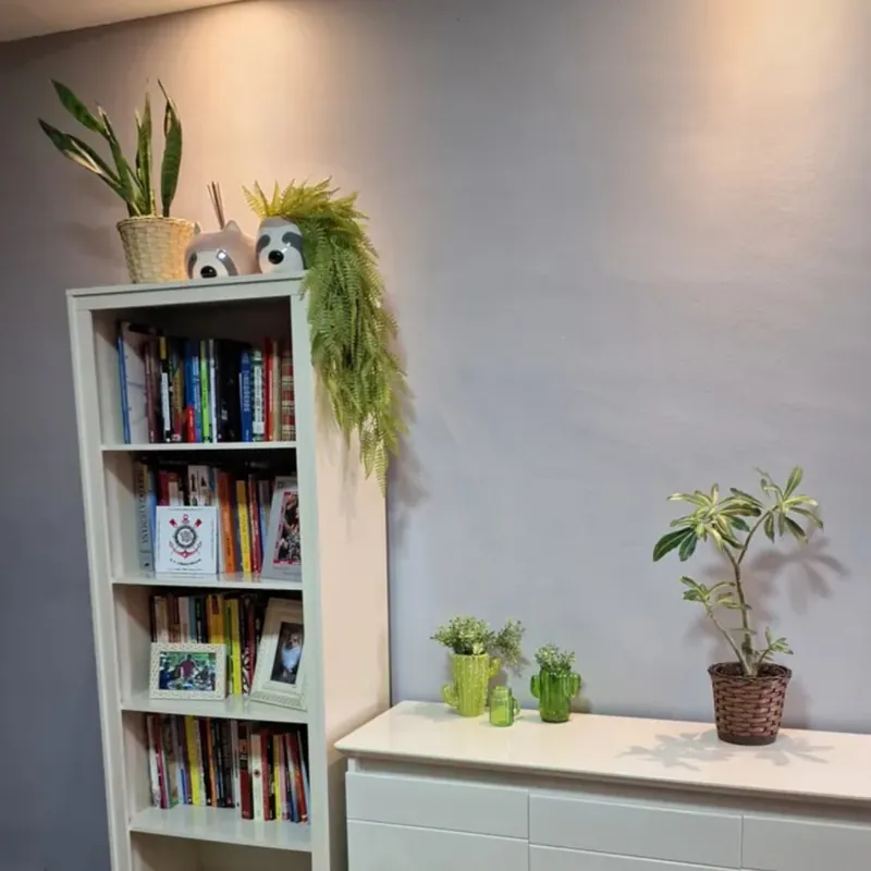 Samambaia artificial verde-clara caindo do alto de uma estante branca com livros, ao lado de vasos e enfeites