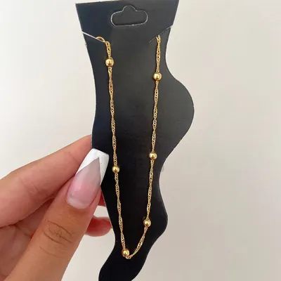Tornozeleira dourada de corrente fina com bolinhas espaçadas, exposta em cartela preta em forma de pé segurada por mão