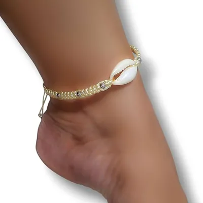 Tornozeleira trançada creme com um búzio branco ao centro e contas prateadas, usada em tornozelo visto de lado
