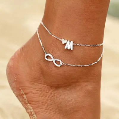 Pé na areia com tornozeleira prateada de duas correntes, uma com coração e letra M, outra com símbolo do infinito
