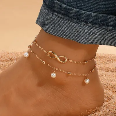 Tornozeleira dourada de duas voltas com infinito e pérolas pendentes, em pé sobre a areia com barra de jeans dobrada