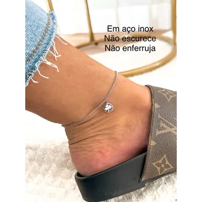 Tornozeleira prateada de corrente fina com medalha redonda de trevo, usada com calça jeans desfiada e chinelo slide