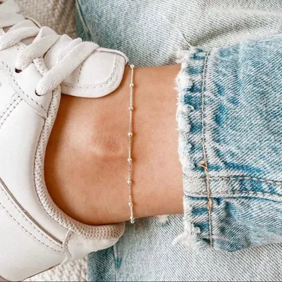 Tornozeleira prateada fina com bolinhas espaçadas no tornozelo, ao lado de um tênis branco e barra de jeans dobrada