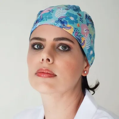 Mulher de jaleco branco usando touca cirúrgica azul-turquesa estampada com folhagens, flores e personagem azul