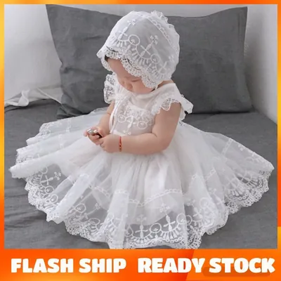 Bebê sentada em cama cinza usando vestido branco de tule bordado com cruzes e touca de renda combinando