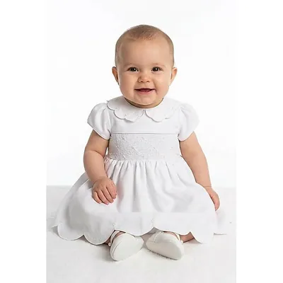 Bebê sentada sorrindo com vestido branco de gola recortada, faixa franzida em casinha de abelha e barra em ondas