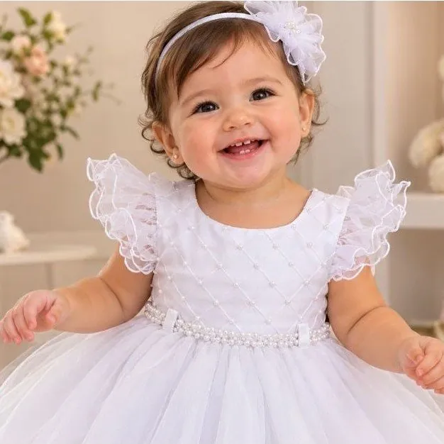 Bebê sorridente com vestido branco de corpinho em losangos com pérolas, mangas de tule em babado e faixa com flor