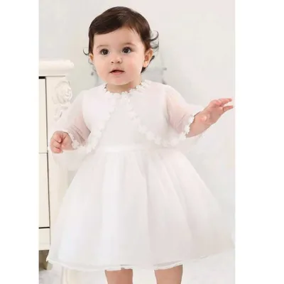 Bebê de vestido branco com saia de tule e xale transparente com flores nas bordas, em quarto claro