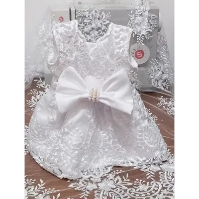 Vestido infantil branco de renda bordada com laço grande de cetim e centro de pérolas, diante de máquina de costura