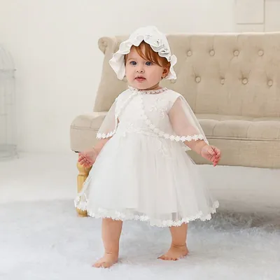 Bebê em pé com vestido branco de tule, capinha transparente bordada e touca de babado, diante de um sofá bege