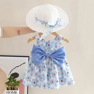 Vestido de bebê branco com flores azuis e laço azul grande, num cabide de madeira, com chapéu branco vazado acima